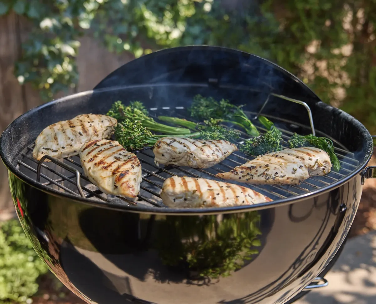 Weber Original Kettle 18" Charcoal Grill (Black), grilling chicken breasts and broccolini in a backyard