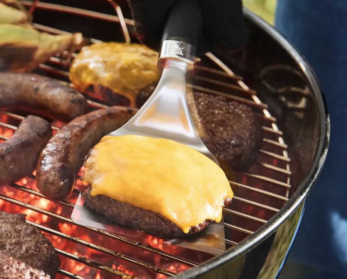 Weber Original Kettle 18" Charcoal Grill (Black), flipping a cheeseburger over hot coals beside sausages